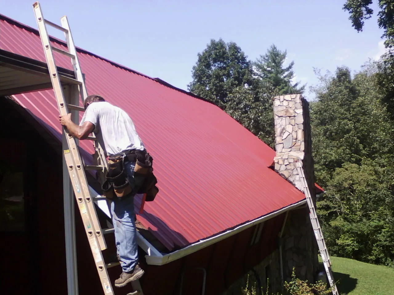 Roofer on a ladder beside a red metal roof and stone chimney — Soffit & Fascia Repair by Roofing Brothers in Trussville